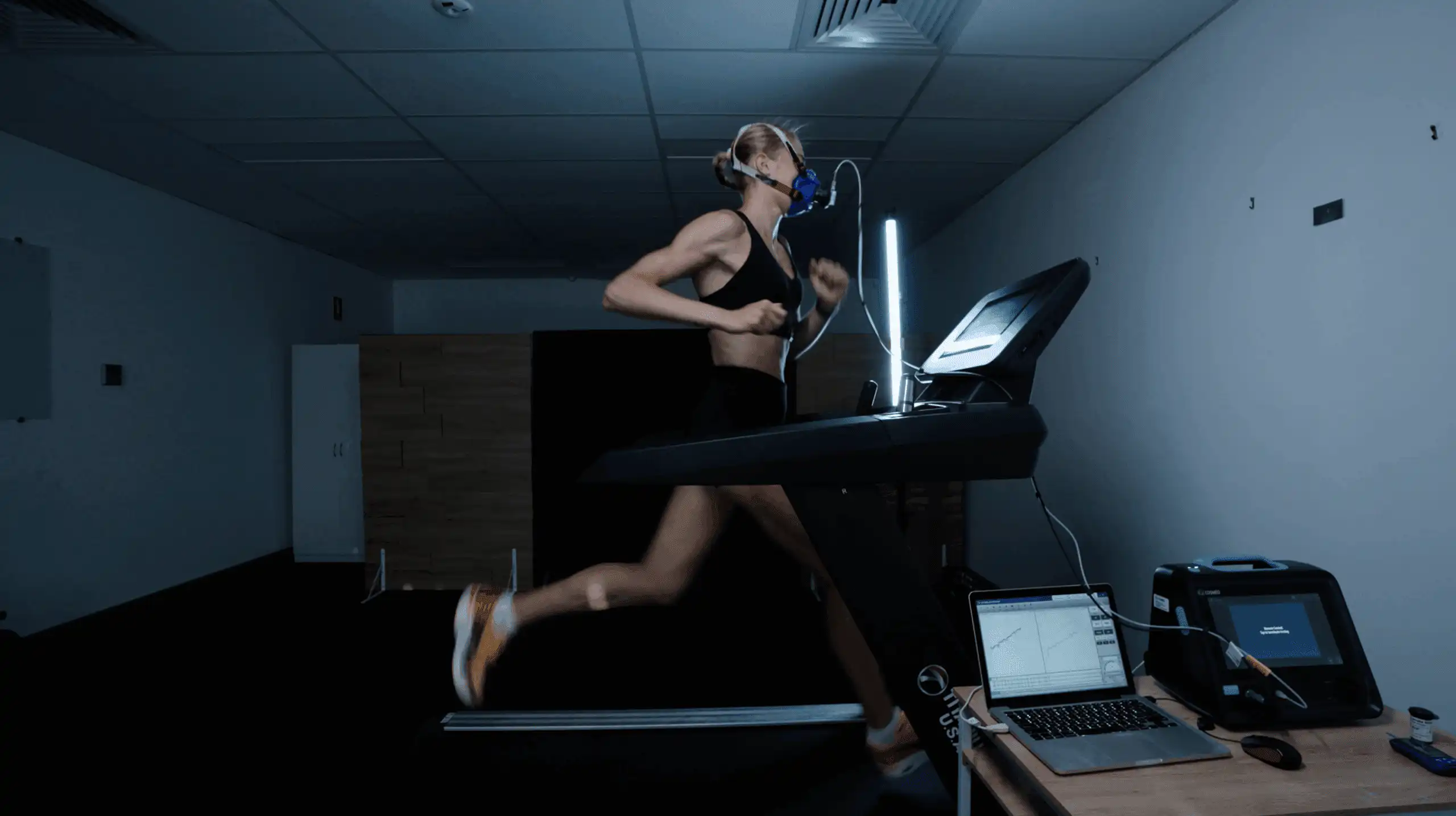 Woman running on a treadmill beside monitoring equipment in a dim room