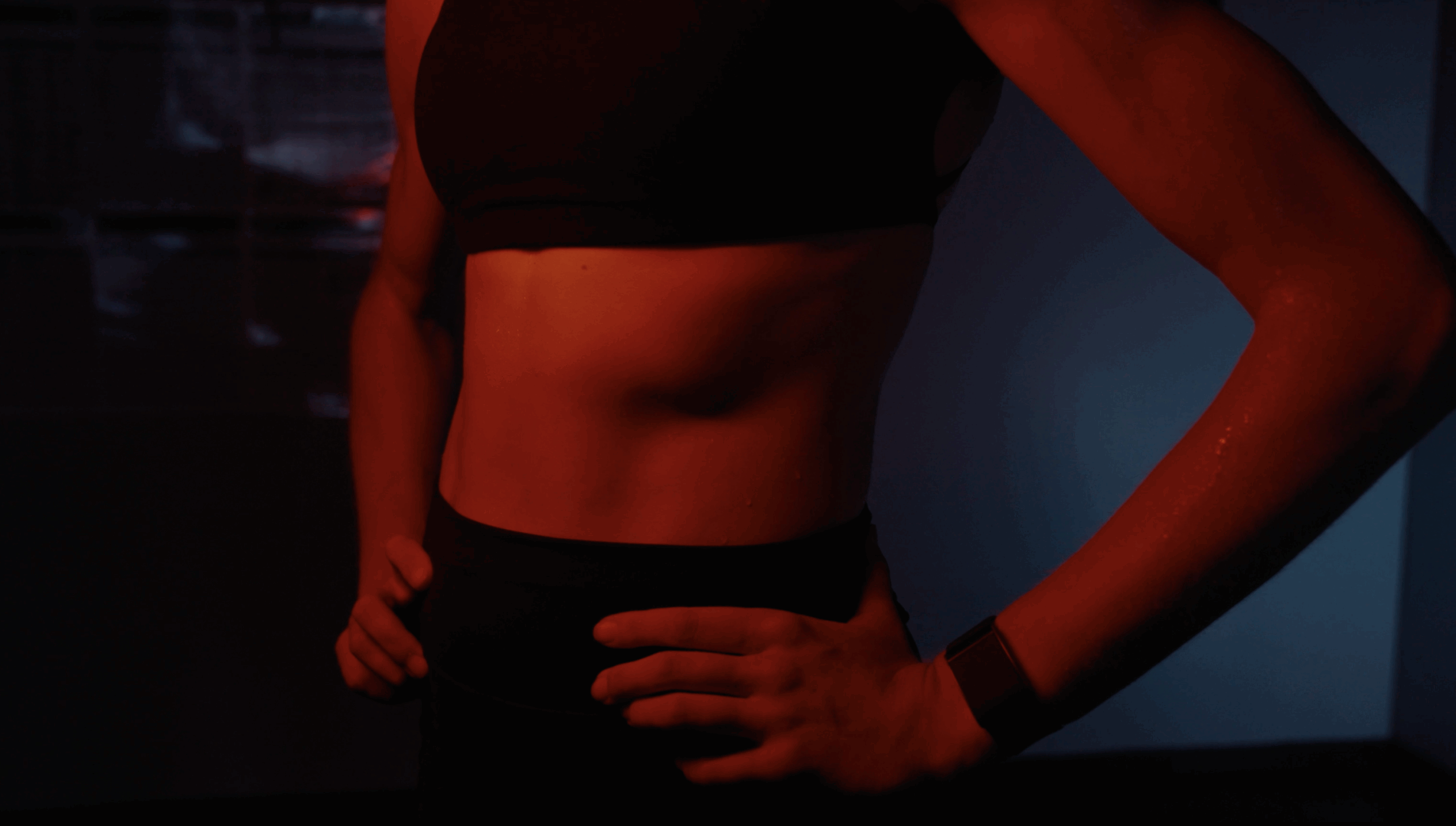 A person wearing a sports bra and fitness watch stands with hands on hips, showing a toned midsection. The lighting is dark with a red and blue tint, creating a dramatic and intense atmosphere.